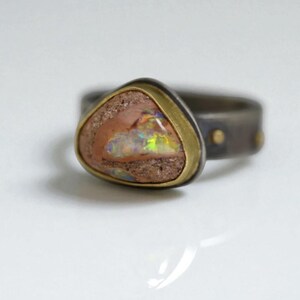 May include: A unique ring featuring a triangular, multi-colored opal stone set in a gold-toned bezel. The band is dark gray with small gold accents. The opal displays a range of colors, including pink, green, and yellow.