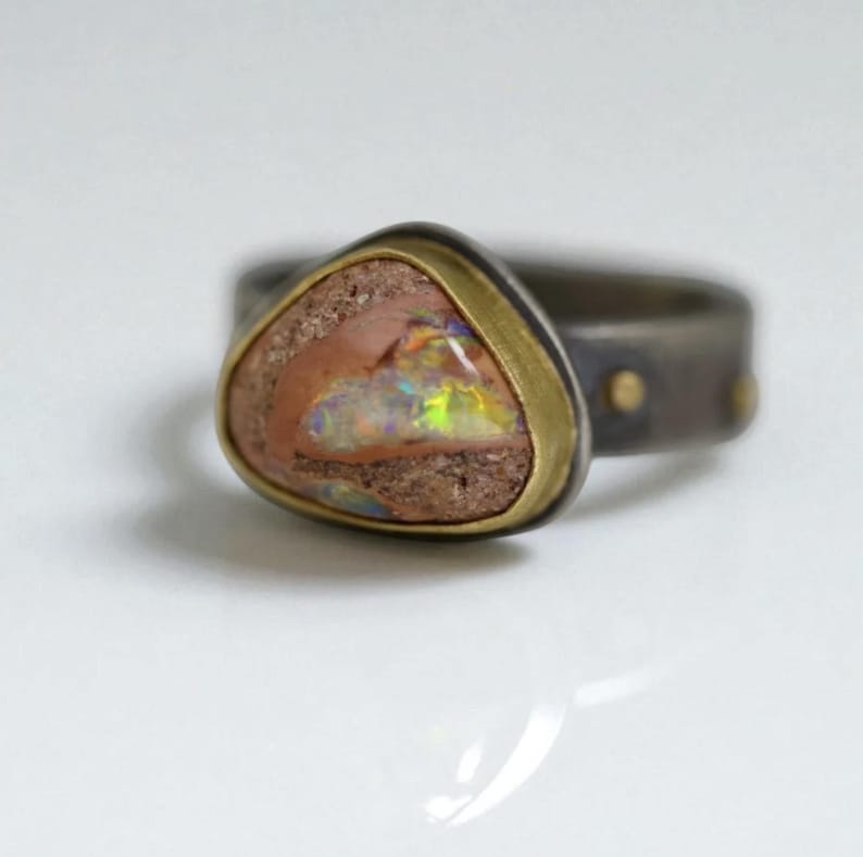May include: A unique ring featuring a triangular, multi-colored opal stone set in a gold-toned bezel. The band is dark gray with small gold accents. The opal displays a range of colors, including pink, green, and yellow.