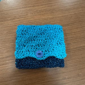 May include: A handmade crochet wallet in shades of blue. The top section is a lighter turquoise, transitioning to a darker navy blue at the bottom. A blue button secures the wallet. The crochet work has a textured, open design.