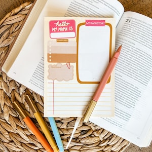 May include: A notepad with the text "hello MY NAME IS" and "MY BACKSTORY" is displayed on an open book. A pink pen and several colorful pens are next to the notepad. The notepad has sections for scriptures, a timeframe, and notes.
