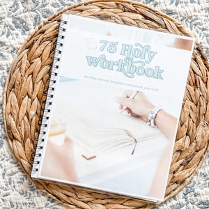 May include: A spiral-bound workbook titled "75 Holy Workbook" with the tagline "develop spiritual disciplines and enrich your faith." The cover features a hand writing in the workbook. The workbook is on a woven, circular placemat.