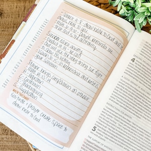 May include: Open Bible with a pink sticky note featuring handwritten notes and Bible verses. The text includes phrases like "draw near to God" and "Prayer brings compassion on others." The page is open to a section with printed text.