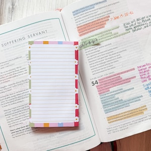 May include: A rectangular notepad with horizontal lines, featuring a white background and a border decorated with small white flowers on a red and green strip. The notepad is placed on an open book with printed text and highlighted passages.