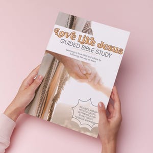 May include: A white and beige book titled "Love Like Jesus Guided Bible Study" is held by two hands against a pink background. The book cover features text and an illustration of hands. The text includes "Learning to love God and others by following the way of Jesus."