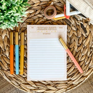 May include: A notepad with the text "Sermon Notes" and lined pages, alongside a pink pen and three colorful pens. The pens are orange, blue, and yellow with gold accents. The items are arranged on a woven, brown surface.