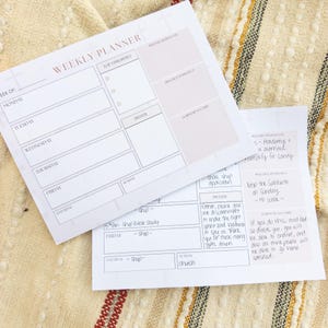 May include: Two white weekly planner sheets with black text and lines. The layout includes sections for days of the week, top priorities, prayer, release or delegate, practice to protect, and scripture cards. One sheet is partially filled with handwritten notes.