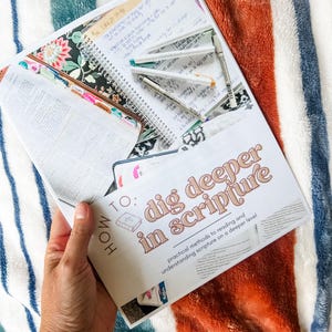 May include: An open book with the text "HOW TO: dig deeper in scripture" and a spiral notebook with handwritten notes and pens. The book has colorful tabs and floral patterned covers. The background is a striped blanket.