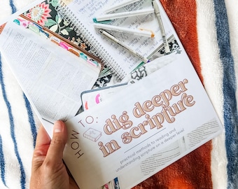 Peut inclure: Un livre ouvert avec le texte "HOW TO: dig deeper in scripture" et un carnet à spirales avec des notes manuscrites et des stylos. Le livre a des onglets colorés et des couvertures à motifs floraux. L'arrière-plan est une couverture rayée.
