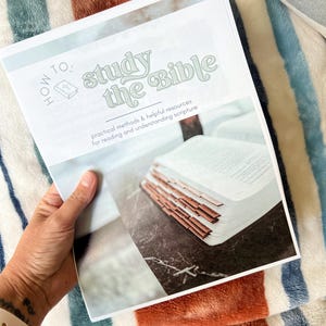 May include: A book titled "How to Study the Bible" with the text in a light green color. The book is open, revealing pages and brown-edged tabs. The background is a patterned blanket with blue, white, and orange stripes.