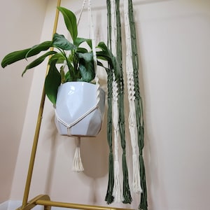 May include: A white ceramic planter with a green plant hanging from a macrame plant hanger. The hanger is made with white and green cotton cord and has a tassel at the bottom.