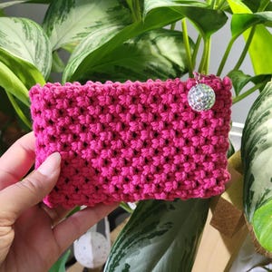 May include: A bright pink, hand-crocheted pouch with a textured pattern. A small, silver disco ball charm hangs from the side. The pouch is rectangular and appears to be made of thick yarn. The background features green plants.