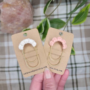 May include: Two handmade paper clips with macrame accents. One clip features a white macrame design, and the other has a pink macrame design. Both clips have gold-colored metal bases and are displayed on brown cardstock.