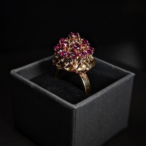 Könnte beinhalten: Ein goldener Ring mit einem Cluster aus rubinroten Steinen, die in einem blumenartigen Design gefasst sind. Der Ring wird in einer schwarzen Samtringbox präsentiert.