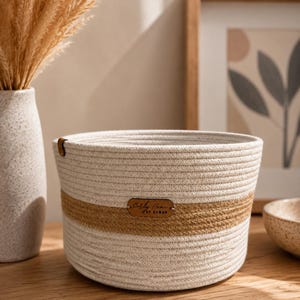 May include: A round, beige rope basket with a brown band around the middle. The basket has a leather tag that reads "Carly Home by SAMAN". The basket is sitting on a wooden surface.