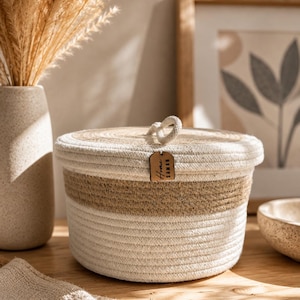May include: A round, lidded storage basket with a natural, woven design. The basket features alternating bands of off-white and tan rope, with a small wooden tag that reads "Home Saman". A loop handle is attached to the lid.