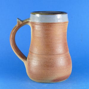May include: A ceramic mug with a textured, reddish-brown body and a smooth, light grey glazed rim. The mug features a curved handle on the left side and a slightly tapered cylindrical shape with visible horizontal ridges along the exterior surface.