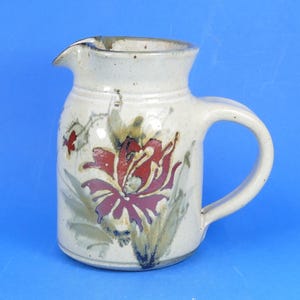 May include: A ceramic jug with a speckled off-white glaze, featuring a hand-painted floral design in shades of deep red, muted green, and brown. The vessel has a wide rim, a curved pouring spout, and a sturdy side handle for easy gripping.
