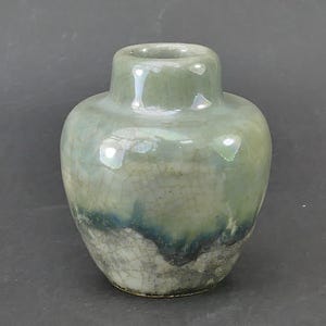 May include: A small ceramic vase with a rounded body and a short, narrow neck. The surface features a glossy, pale green glaze with a visible crackle pattern throughout, transitioning to a darker blue and grey textured base at the bottom.