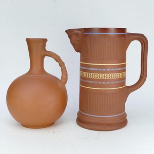 May include: A terracotta-coloured ceramic jug with a rounded body and a textured handle alongside a cylindrical terracotta pitcher featuring a ram's head detail, vertical ribbed bands, and horizontal stripes in light blue and yellow.