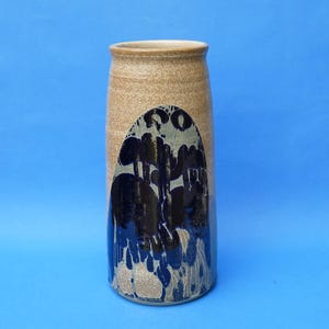 May include: A cylindrical ceramic vase with a speckled tan base and a prominent, arched decorative panel featuring dark blue and black glaze that drips downwards, creating a textured, layered appearance against the lighter clay body.