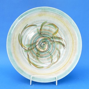 May include: A round ceramic bowl featuring a hand-painted spiral design in shades of sage green, soft yellow, and earthy brown. The interior surface displays concentric circular ridges with a glossy finish, radiating from the centre outwards.