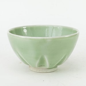 May include: A small, light green ceramic bowl with a glossy finish and a fluted, petal-like exterior design. The bowl features a rounded shape that tapers down to a plain, unglazed circular base, showcasing a smooth, uniform pale green glaze.