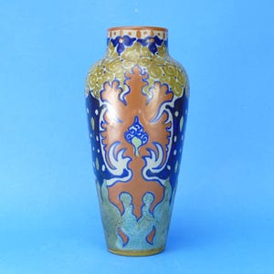 May include: A ceramic vase featuring a symmetrical, ornate pattern in shades of terracotta, deep blue, and muted gold. The design includes stylised floral and scroll motifs that wrap around the body, with a smooth, tapered neck and a wide base.
