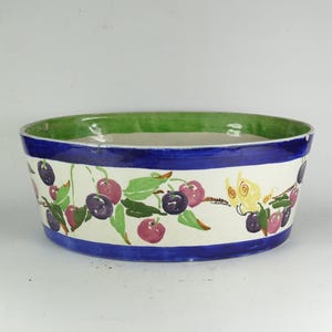 May include: Oval ceramic bowl with a white exterior, featuring a blue rim and a green interior rim. The bowl is decorated with a cherry branch design in shades of red, purple, and green.