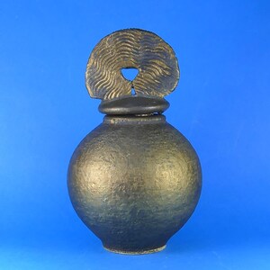 May include: A dark bronze-coloured ceramic vessel with a spherical body and a textured surface. The vessel features a lid topped with a semi-circular handle that has a wavy, radial pattern and a small central opening, creating a distinct silhouette.