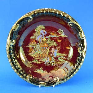 May include: A decorative maroon plate with a gold and black ornate rim. The centre features a detailed landscape scene with buildings, trees, and figures in gold, green, and blue. The plate is displayed on a white stand.