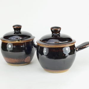 May include: Two dark brown ceramic pots with lids. One pot has a handle. The pots have a glossy finish and a light brown rim. The pots are likely for cooking or serving food.