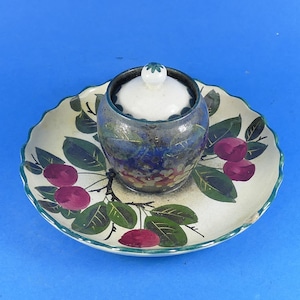 May include: A ceramic lidded jar sits centred on a matching scalloped plate. Both pieces feature a cream-coloured base with a painted design of dark green leaves and round purple fruit. The jar has a white lid with a small green floral knob.