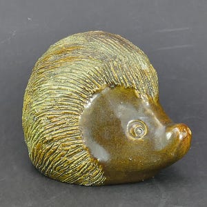 May include: A ceramic hedgehog figurine featuring a textured, golden-brown back that mimics quills and a smooth, dark brown face. The sculpture has a glossy finish, a pointed snout, and a single visible eye, capturing the animal in a curled position.