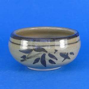 May include: A round ceramic bowl with a glossy beige glaze, featuring a dark brown horizontal band near the rim and a hand-painted dark brown leaf motif encircling the exterior. The interior of the bowl is a smooth, light cream colour.