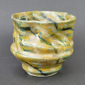 May include: A ceramic cup with a wavy, irregular rim and a textured, hand-formed body. The surface features a marbled glaze pattern in shades of mustard yellow, deep teal, and off-white, creating a dynamic, layered appearance across the exterior.