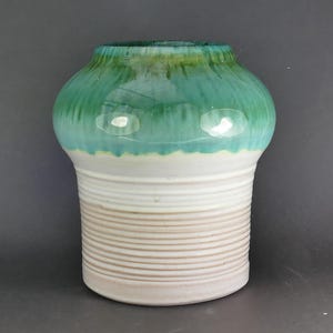 May include: A ceramic vase featuring a wide, rounded upper section with a glossy, dripping emerald green glaze that transitions into a smooth, white middle. The lower portion is cylindrical with horizontal ribbed textures in a light beige tone.