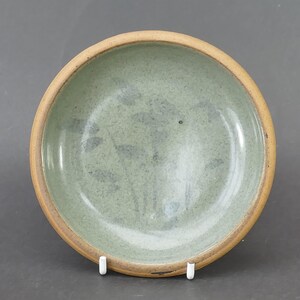 May include: A round ceramic bowl featuring a speckled light green interior glaze with a subtle dark leaf pattern. The bowl has a smooth, unglazed tan rim that contrasts with the glazed centre, creating a simple and functional kitchen vessel.