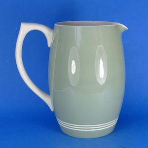 May include: A pale green ceramic jug with a white handle and spout. The jug features two white oval reflections and two white stripes near the base. The handle is a curved, open design. The jug is a classic shape.