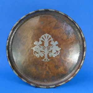 May include: A round copper tray featuring a hammered texture and a central silver-toned floral inlay. The rim is decorated with an alternating pattern of copper and silver-toned segments, creating a distinct border around the dark metallic surface.