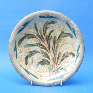 May include: A round ceramic bowl featuring a hand-painted botanical design with dark green and brown leafy sprigs. The interior surface has a mottled beige and cream background with thin, swirling brown lines and teal accents along the rim.