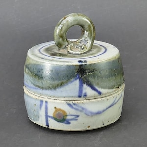 May include: A cylindrical ceramic lidded container with a loop handle on top. The piece features a speckled off-white glaze with hand-painted blue linear accents, a green wash across the upper section, and a small circular orange and green motif.