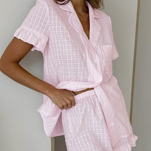 Puede incluir: Un conjunto de pijama rosa claro con un patrón geométrico texturizado. La parte superior de manga corta tiene cuello, botones y un bolsillo en el pecho. Los pantalones cortos tienen un dobladillo con volantes. La tela parece ligera y cómoda.