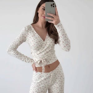 以下が含まれることがあります： クリーム色のフローラルプリントのラップトップスと、お揃いのパンツ。長袖のトップスはVネックで、ウエストにタイクロージャーが付いています。パンツはワイドレッグです。フローラルパターンは小さなピンクの花が特徴です。