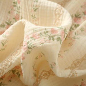May include: Close-up of a cream-colored fabric with a delicate floral pattern. Pink roses and green leaves are scattered across the material, with a decorative border in a muted brown tone. The fabric appears soft and lightweight.