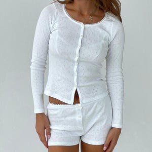 Könnte beinhalten: Weißes, langärmliges Oberteil mit Knöpfen und passendem Shorts-Set. Das Oberteil hat einen runden Ausschnitt mit Spitzenbesatz und einem kleinen Punktmuster. Die Shorts haben ebenfalls einen Spitzenbesatz und einen Knopfverschluss.