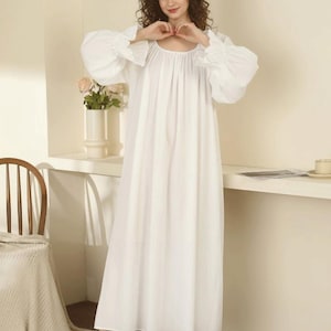 May include: A long, white nightgown with voluminous sleeves and a gathered neckline. The gown is floor-length and paired with white, fluffy slippers. A wooden chair and a vase of flowers are visible in the background.
