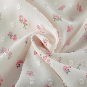 Puede incluir: Primer plano de una tela color crema con un delicado estampado floral. La tela presenta peque&ntilde;os puntos en relieve y est&aacute; adornada con ilustraciones de rosas rosas y verdes. La tela est&aacute; drapeada, mostrando su textura y dise&ntilde;o.