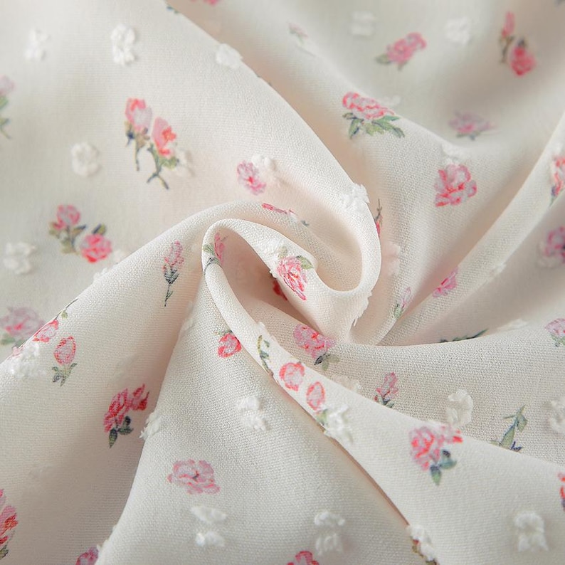 Puede incluir: Primer plano de una tela color crema con un delicado estampado floral. La tela presenta peque&ntilde;os puntos en relieve y est&aacute; adornada con ilustraciones de rosas rosas y verdes. La tela est&aacute; drapeada, mostrando su textura y dise&ntilde;o.