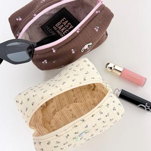 May include: Two floral print cosmetic bags, one brown with pink accents and the other cream with blue accents, are open to reveal their interiors. A pair of sunglasses and makeup items are placed near the bags.
