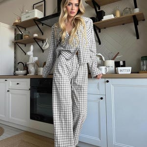 May include: A matching pajama set in a gray and white checkered pattern. The set includes a long-sleeved button-up shirt and wide-leg pants. The outfit is made of a soft, lightweight fabric.